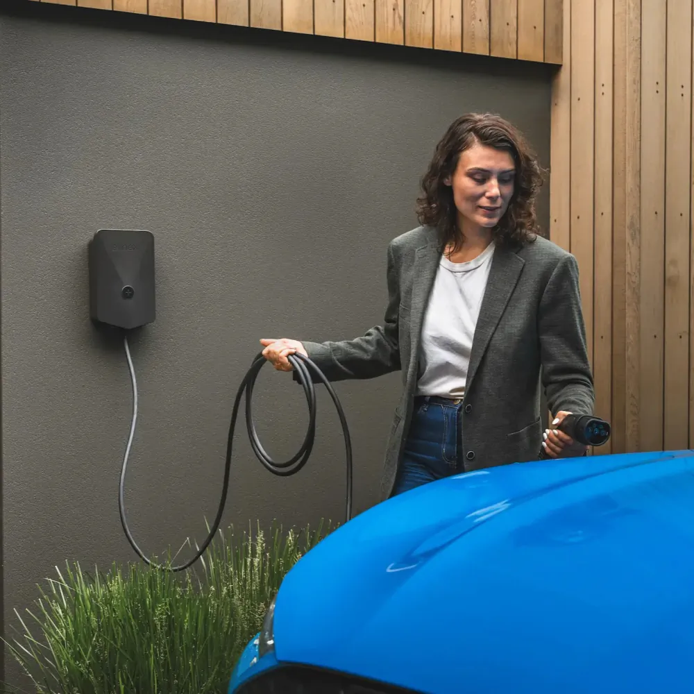 Lifestyle shot of a women using a wall-mounted Evnex E2 Flex EV charger to plug into a bright blue electric vehicle.